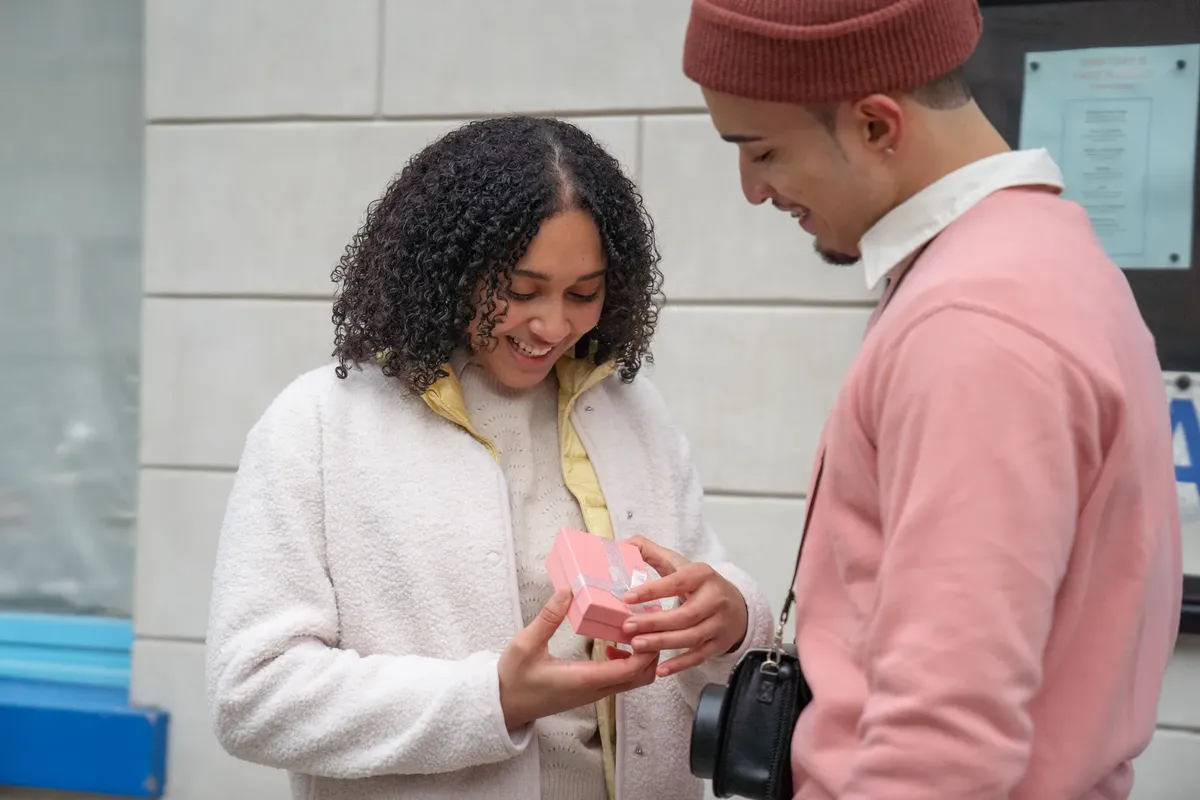 Smiling Hispanic girlfriend with pink gift box standing near positive boyfriend on street near building during romantic date in city