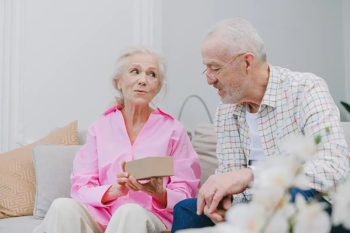 Elderly couple sitting indoors, sharing a heartfelt moment with a gift.